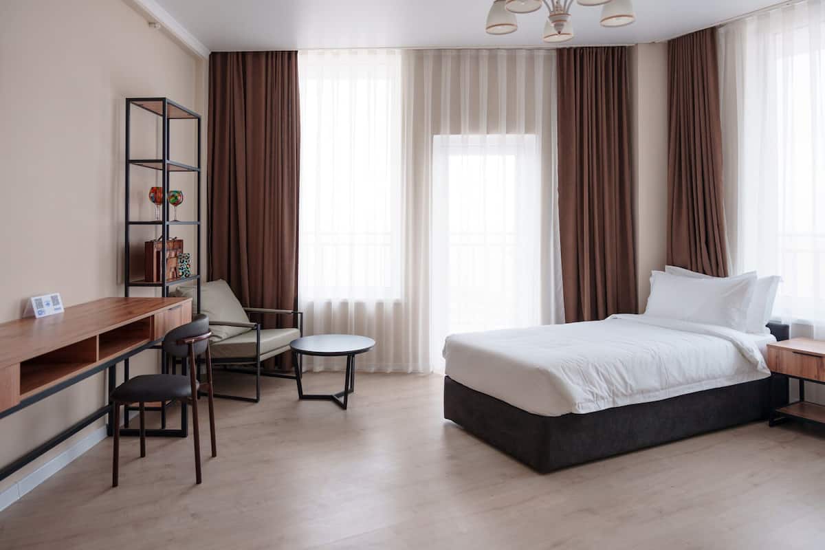 A hotel room with a bed, a desk, a chair, a small table, and a bookshelf.