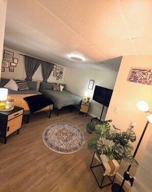 Ein Schlafzimmer mit einem Bett, Nachttischen, einer Lampe, einer Stehlampe, einer Topfpflanze und einem gemusterten Teppich.