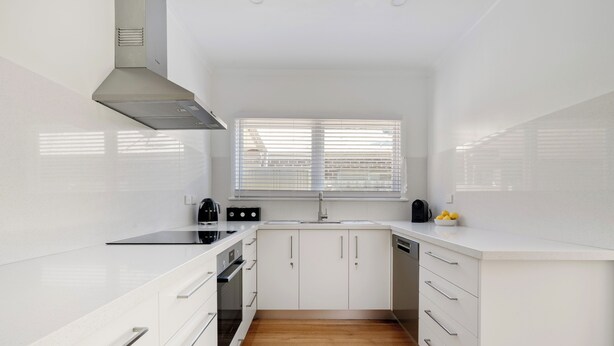 Eine moderne Küche mit weißen Schränken, einer Edelstahl-Dunstabzugshaube, einem eingebauten Backofen, einem Geschirrspüler und einem Fenster mit Jalousien.