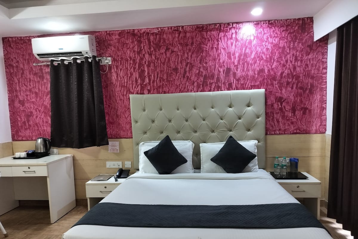 A hotel room with a large bed, a bedside table, a nightstand, a wall-mounted air conditioner, and a decorative red wall.