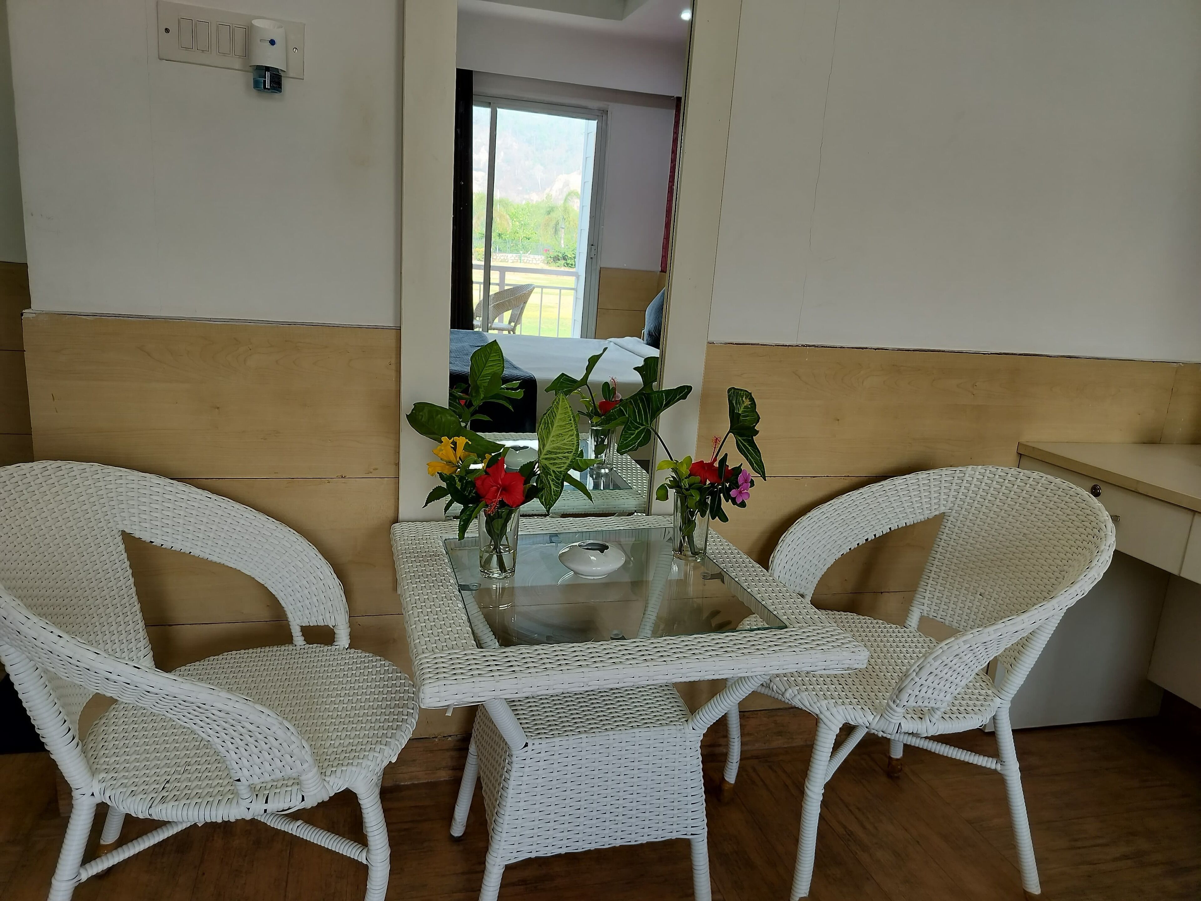 Una habitación con una mesa de vidrio, dos sillas blancas de mimbre y una mesita con un jarrón de flores.