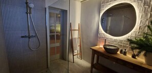 Un baño con cabina de ducha de vidrio, tocador de madera con espejo redondo y una planta en maceta.