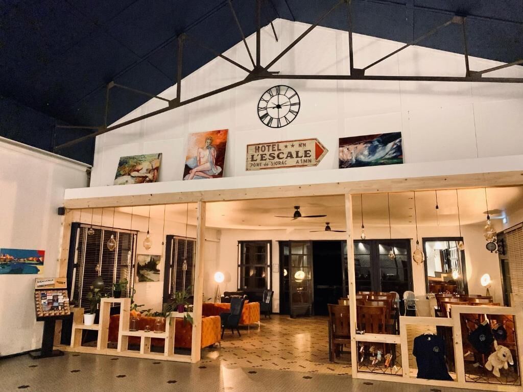 A hotel entrance with a clock, a sign reading "HOTEL L'ESCALA," and a view of the interior including seating areas and a dining space.