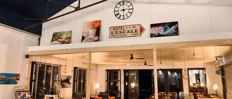 A hotel entrance with a clock, a sign reading "HOTEL L'ESCALA," and a view of the interior including seating areas and a dining space.