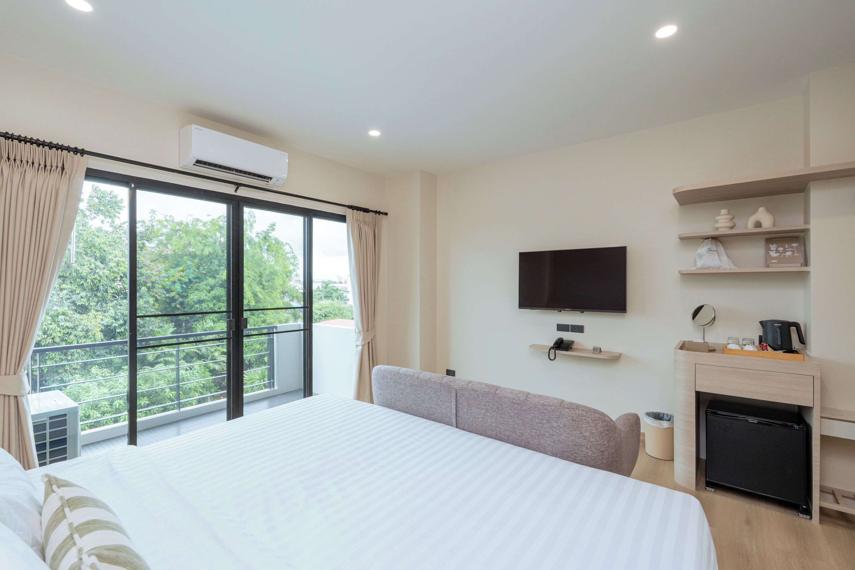 A hotel room with a large bed, a TV mounted on the wall, a small shelf with decorative items, and a view of greenery through the window.