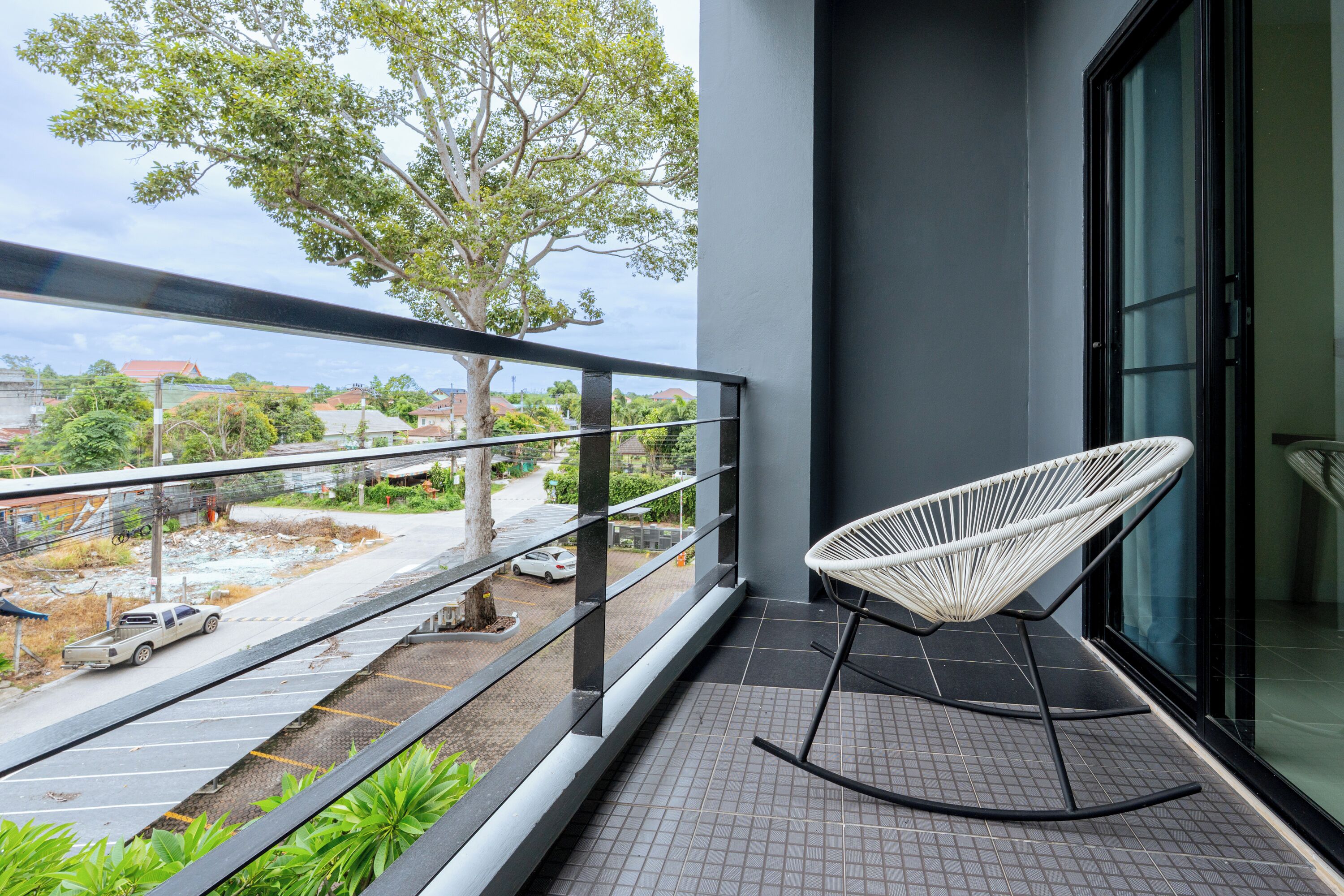 A balcony with a white woven chair, a metal railing, and a view of a street with parked cars and trees.