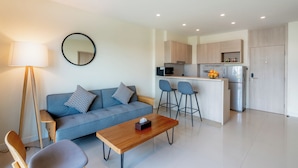 A modern living room with a sofa, coffee table, and bar stools. A kitchen with a refrigerator and microwave is visible in the background. - Aster Residence Rayong (Rayong)