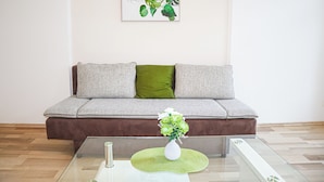 Een moderne woonkamer met een grijze bank, groen- en grijze kussens, een glazen salontafel met een witte vaas, en een kunstwerk boven de bank.