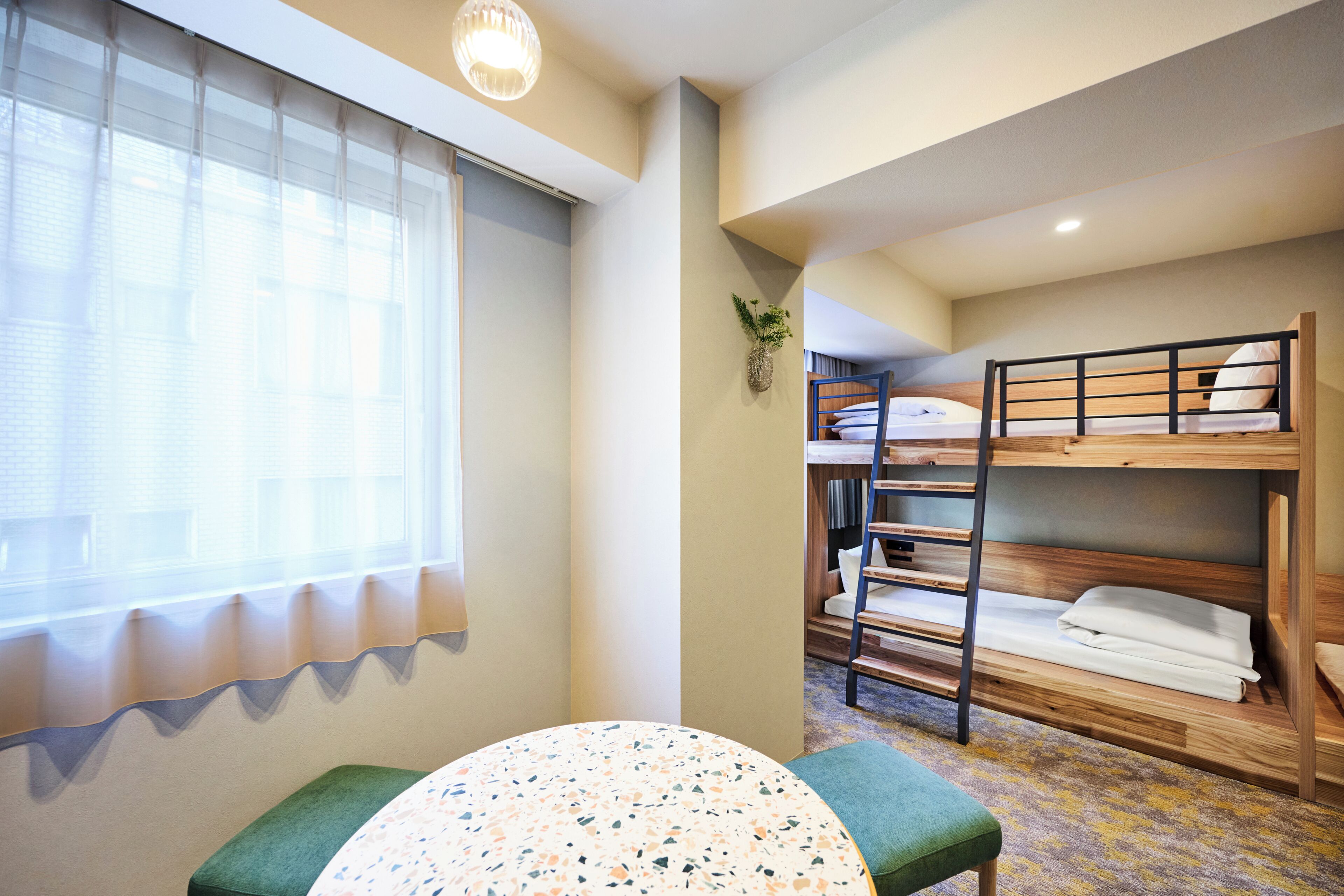 Quarto com beliche, mesa e banco, janela com persianas e uma planta na parede.