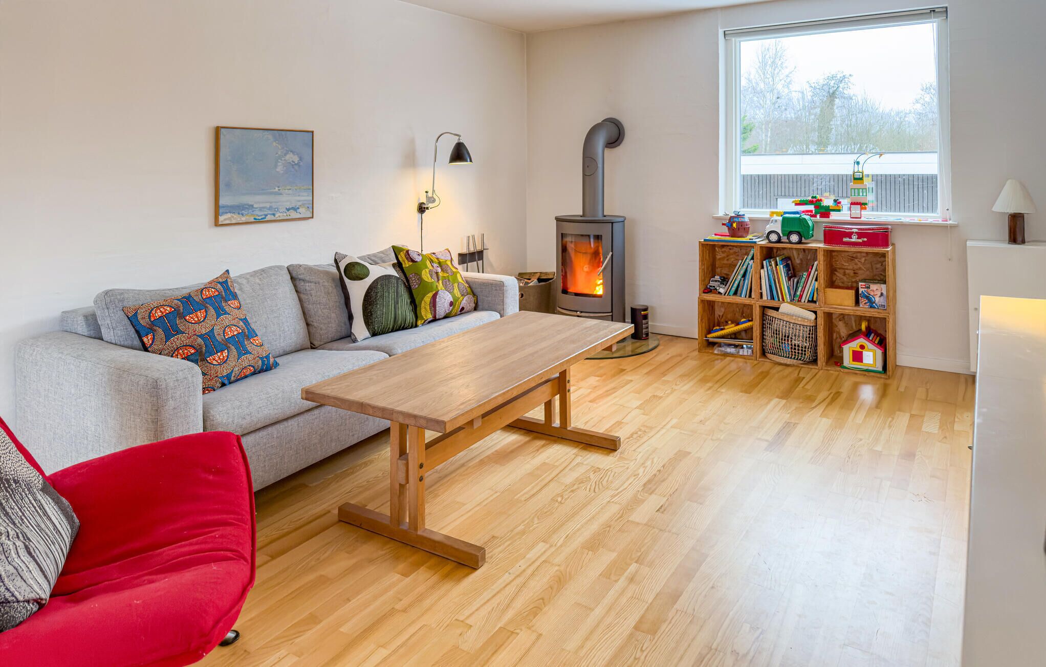 Ein gemütliches Wohnzimmer mit einer Couch, einem Holztisch, einem roten Sessel, einem Bücherregal mit Spielzeug und einem Kamin.