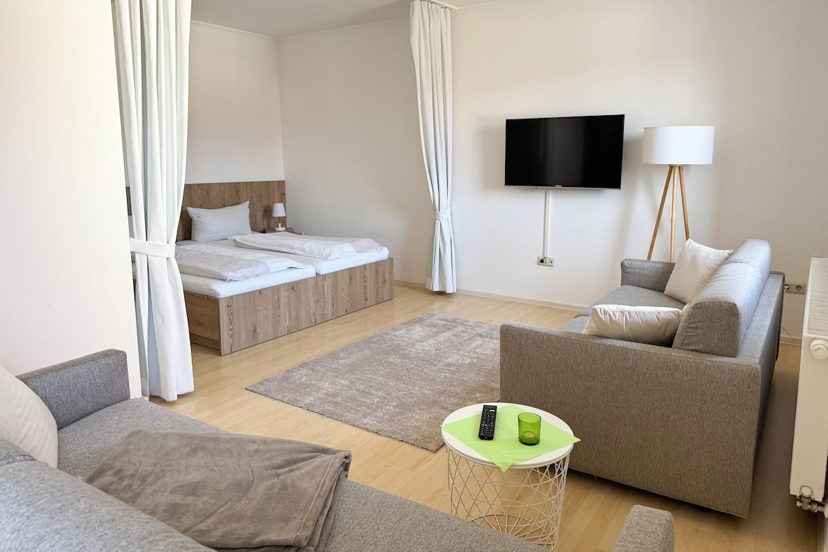 Moderni hotellihuone, jossa on sänky, sohva, pieni pyöreä pöytä, jossa on kaukosäädin ja lasi, sekä seinälle asennettu televisio.
