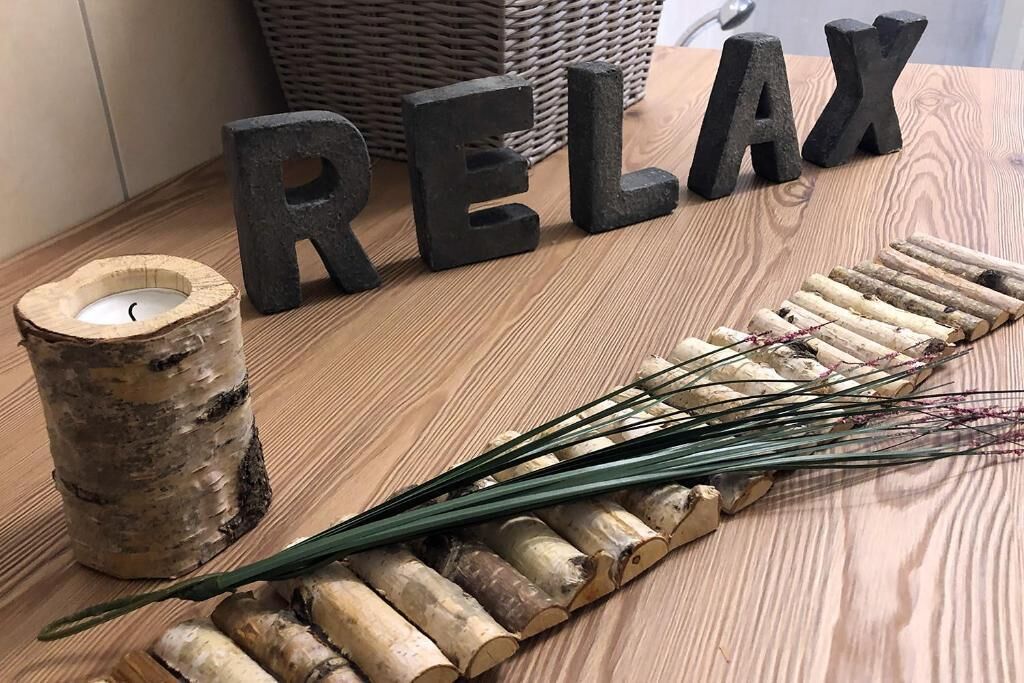 A wooden table with the word "RELAX" spelled out in black letters, a candle in a birch log holder, and a row of dried reeds.