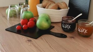 Une table en bois sur laquelle se trouvent un saladier de pommes, un panier de pain, une bouteille de jus d’orange et deux pots de pâte à tartiner au chocolat de la marque Gluck.