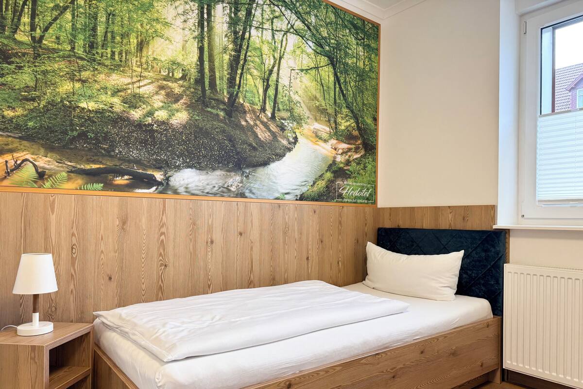 Makuuhuoneessa on puinen sängynpääty, valkoisilla tekstiileillä varustettu sänky, yöpöytä lampun kanssa ja suuri seinämaalaus, jonka aiheena on metsä.