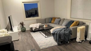 Un salon moderne avec un canapé gris, une table basse et un radiateur. On aperçoit une fenêtre donnant sur des arbres et une chaise à l’extérieur.