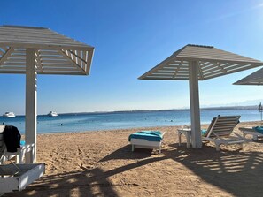 Ein Strand mit weißen Sonnenschirmen und Liegestühlen, feinem Sand und Booten im Wasser.