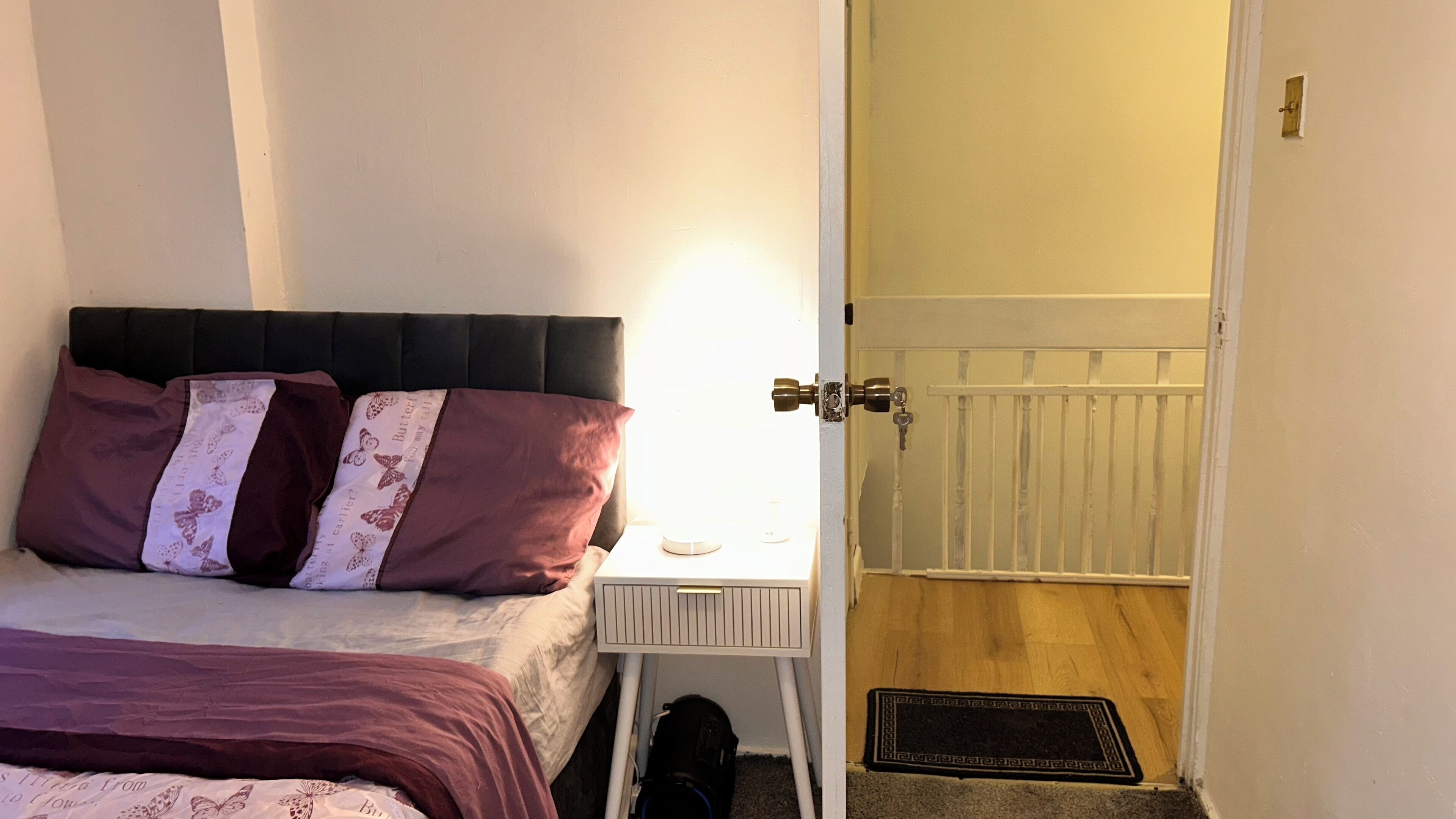 Un dormitorio con cama, almohadas, mesita de noche, lámpara, radiador y puerta.