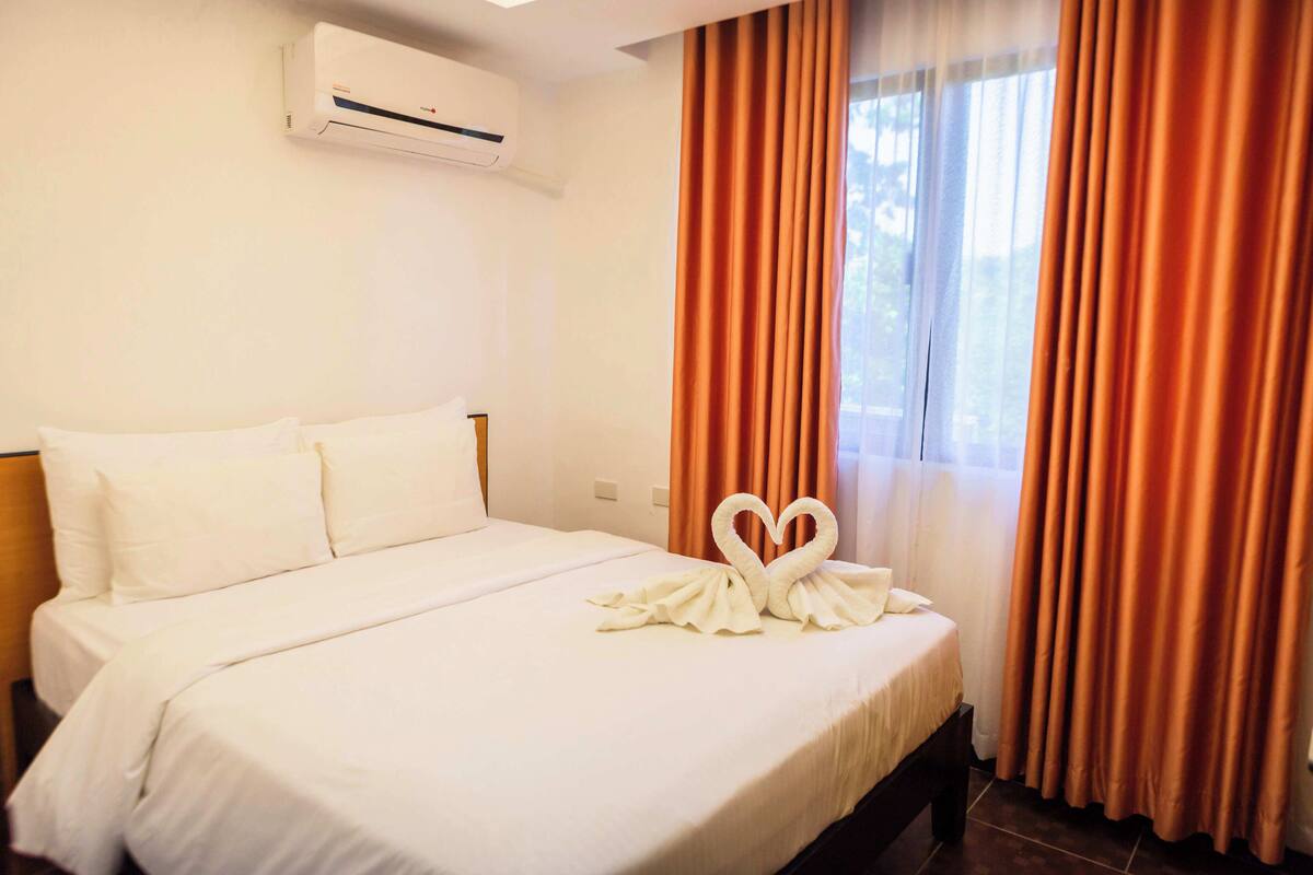 A hotel room with a neatly made bed, an air conditioning unit on the wall, and orange curtains.