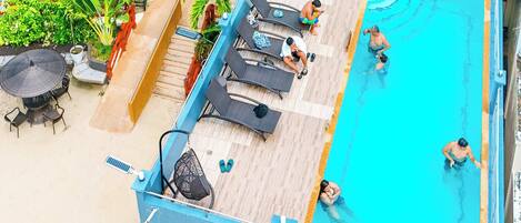 Un área de piscina en la azotea con sillones de descanso, una mesita y algunas personas nadando y relajándose.