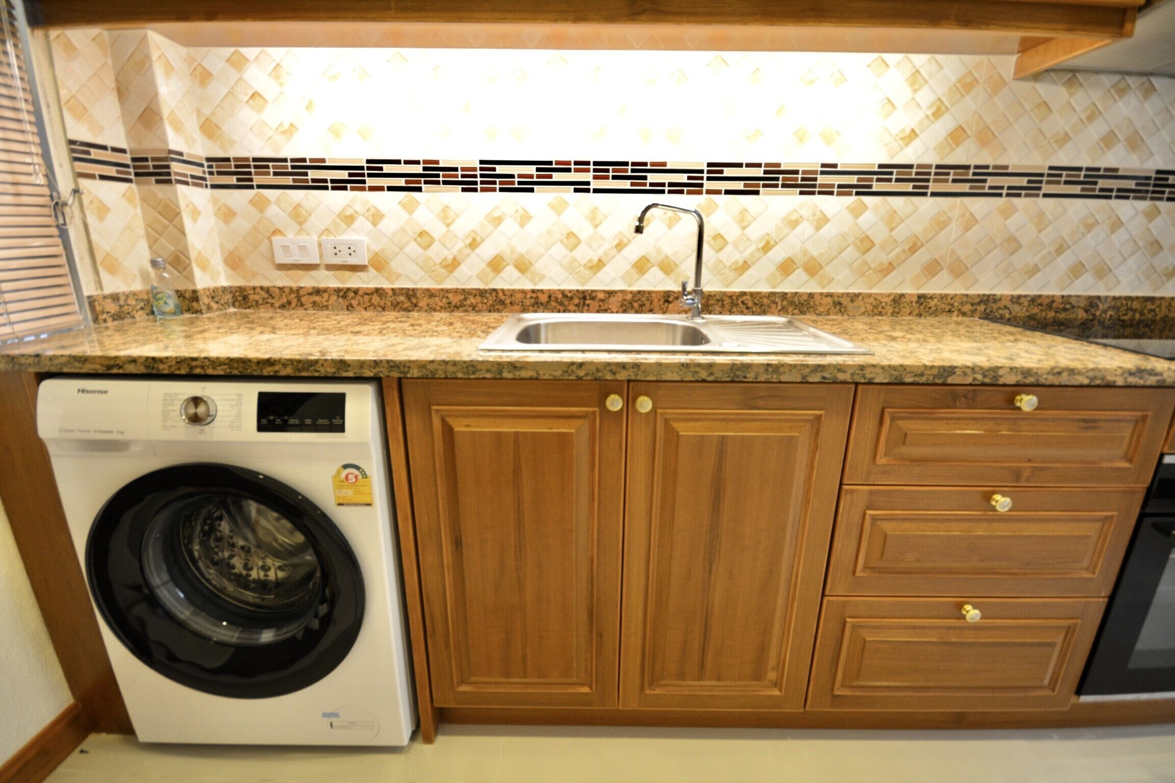 A compact kitchen with a built-in washing machine, wooden cabinets, a granite countertop, and a tiled backsplash.