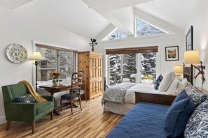 A cozy bedroom with a wooden floor, a green armchair, a bed with blue bedding, and a window with a view of snow-covered trees. - Ski-Access Studio in Mountain Village | Blue Mesa Lodge 33C by AvantStay (Telluride)