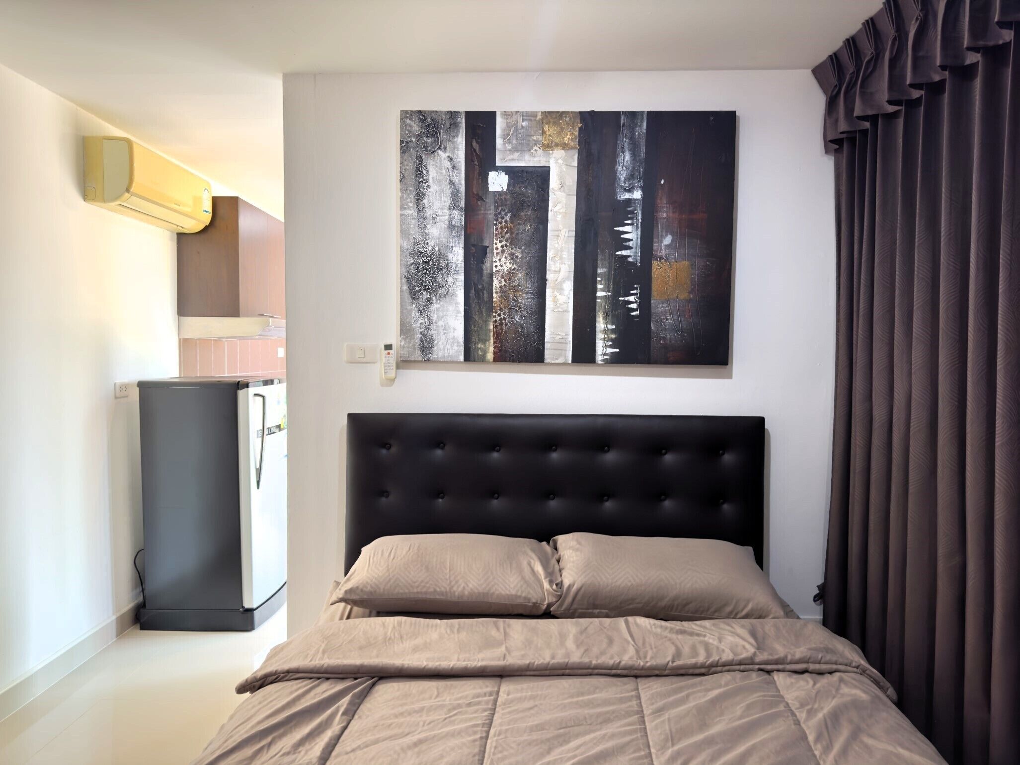 A bedroom with a bed, a wall-mounted air conditioner, a refrigerator, and a painting.
