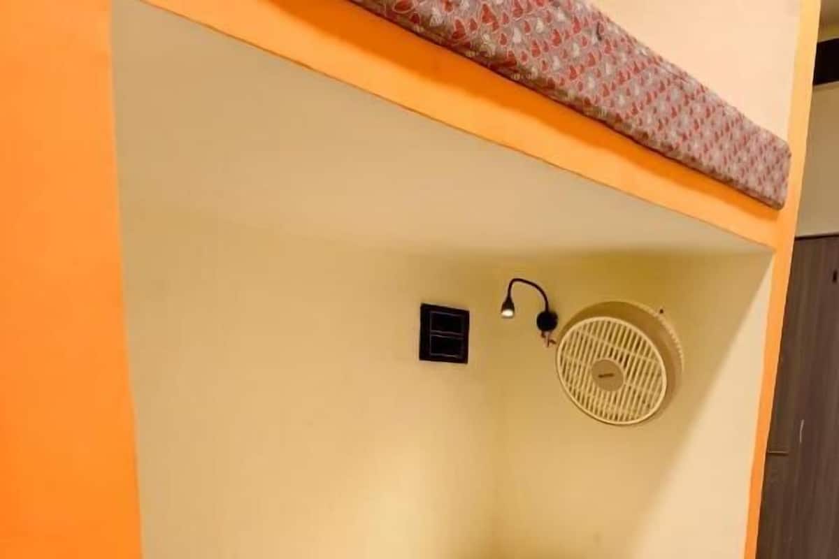 Ein Etagenbett mit gemustertem Bettbezug, einem weißen Kopfkissen, einem kleinen schwarzen Gegenstand an der Wand und einem Wandventilator.