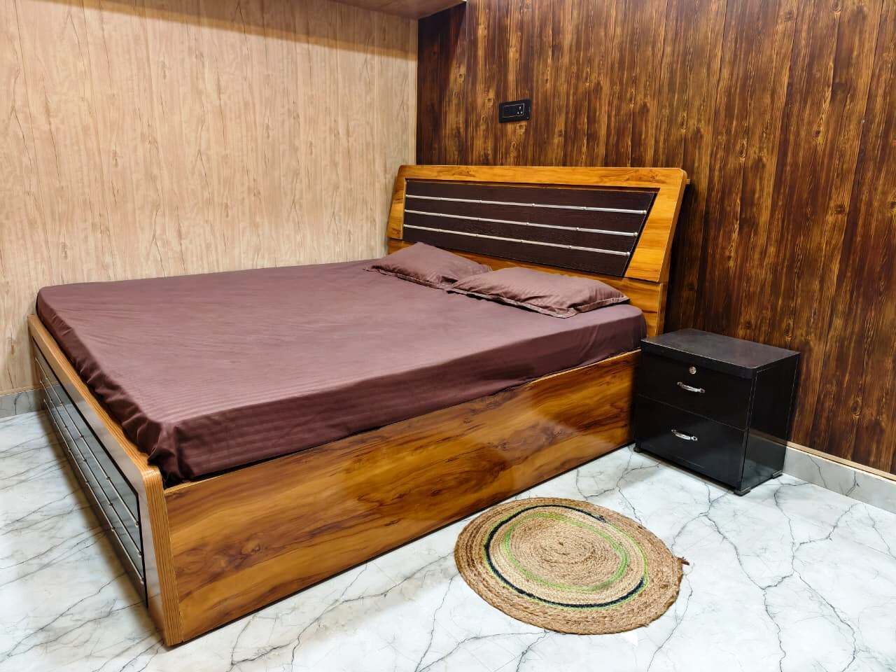 A wooden bed with a headboard, a small black nightstand, and a woven rug on a marble floor.