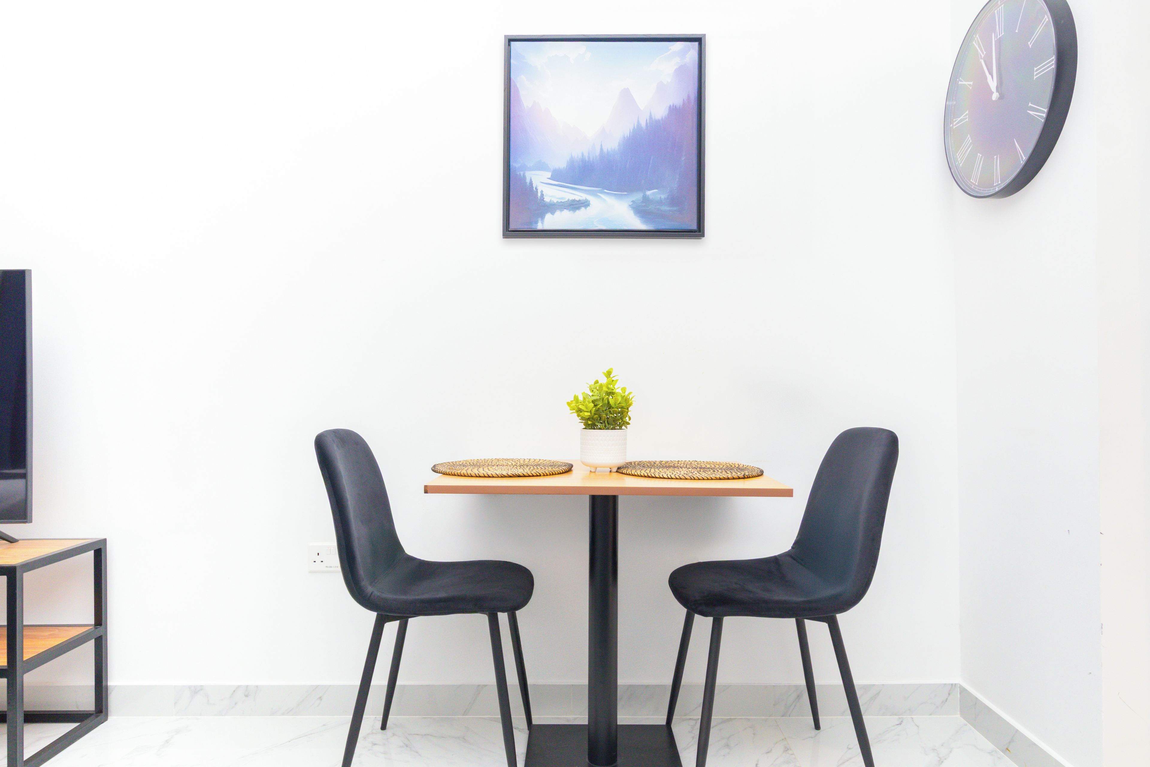 Uma sala de jantar moderna com mesa de madeira, duas cadeiras pretas, um vaso com planta e um quadro de paisagem na parede.