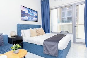 A modern bedroom with a bed, a nightstand, a lamp, a chair, and a window with curtains. - Bloomfields Studio in Oasis 1-Masdar (Abu Dhabi)