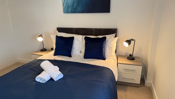 Una cama bien tendida con colcha azul oscuro, dos almohadas blancas y dos almohadas azul oscuro. Hay dos mesitas de noche con lámparas, una obra de arte colgada en la pared y un suelo alfombrado.