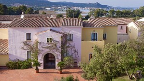 Un patio con edificios que tienen techos de teja y paredes de colores pastel, rodeado de vegetación y una vista distante de un pueblo y montañas.