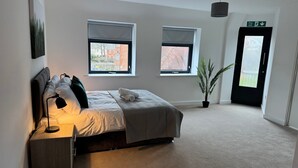 Quarto com cama, abajur, janelas, planta em vaso e porta com placa de saída de incêndio.