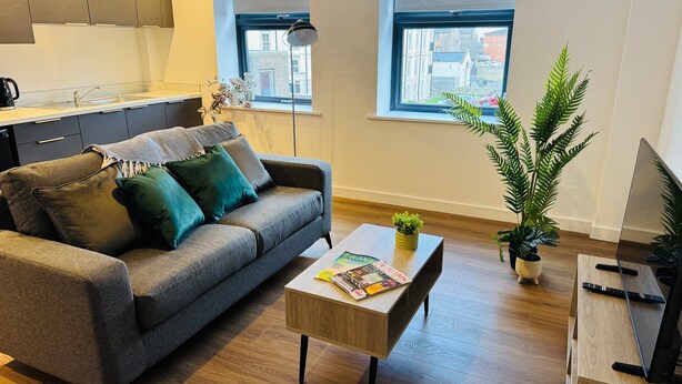 Uma sala de estar moderna com um sofá cinzento, uma mesa de centro com revistas, uma planta em vaso e vista da cidade através de grandes janelas.