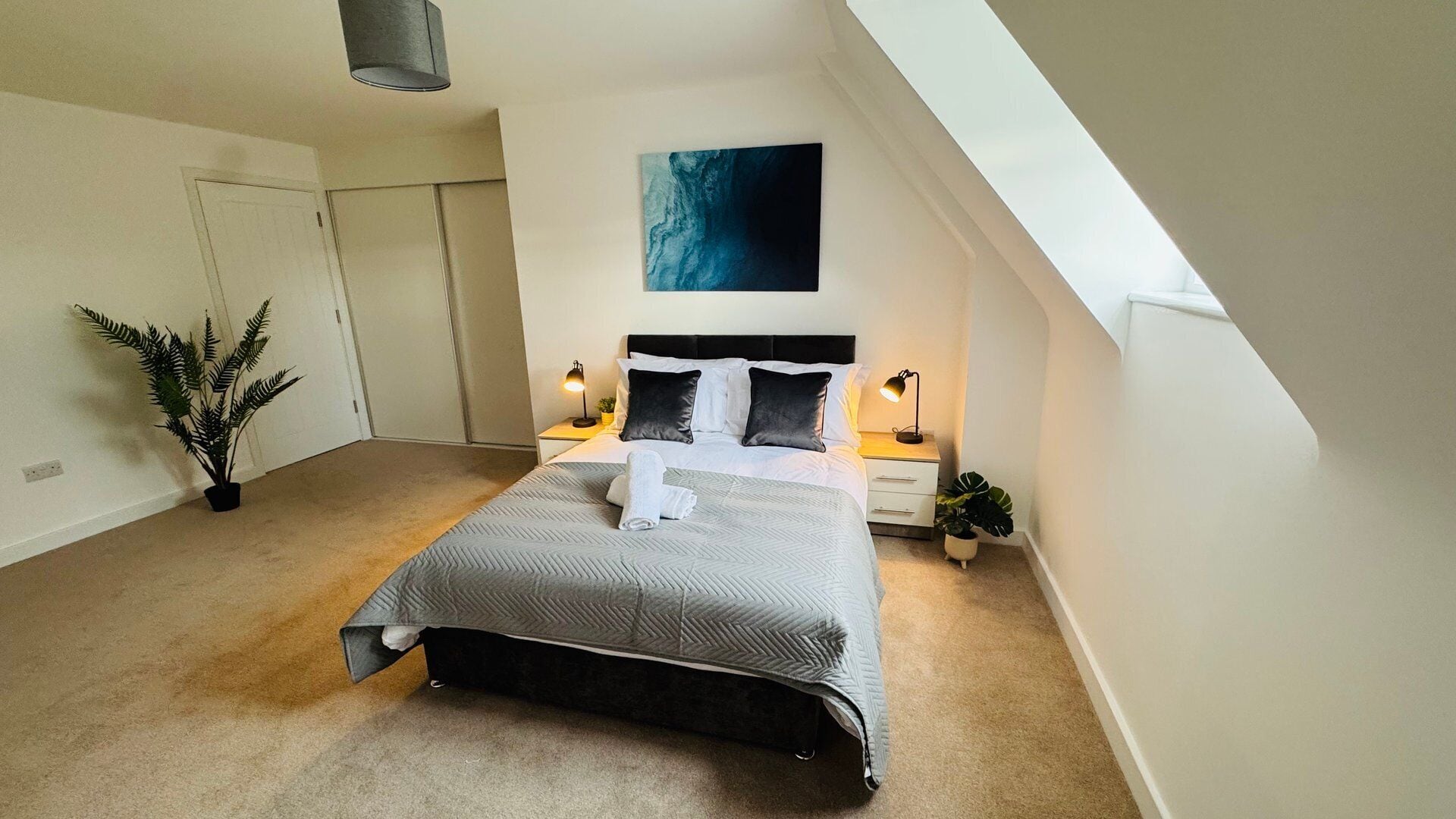A bedroom with a bed, bedside lamps, a potted plant, and a painting on the wall.
