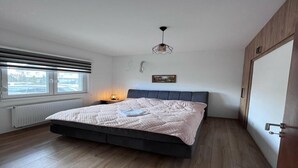 A bedroom with a bed, a bedside lamp, a window with blinds, a picture on the wall, and a door.