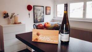 Une bouteille de vin, une boîte en bois et un panier de fruits sont posés sur une table dans une pièce dotée d’une fenêtre et ornée d’œuvres d’art au mur.