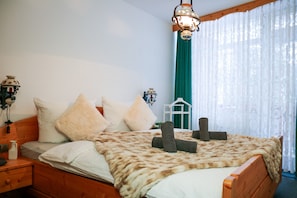 Un dormitorio con cama de madera, almohadas blancas y beige, manta tipo piel, lámpara colgante y ventana con cortinas traslúcidas.