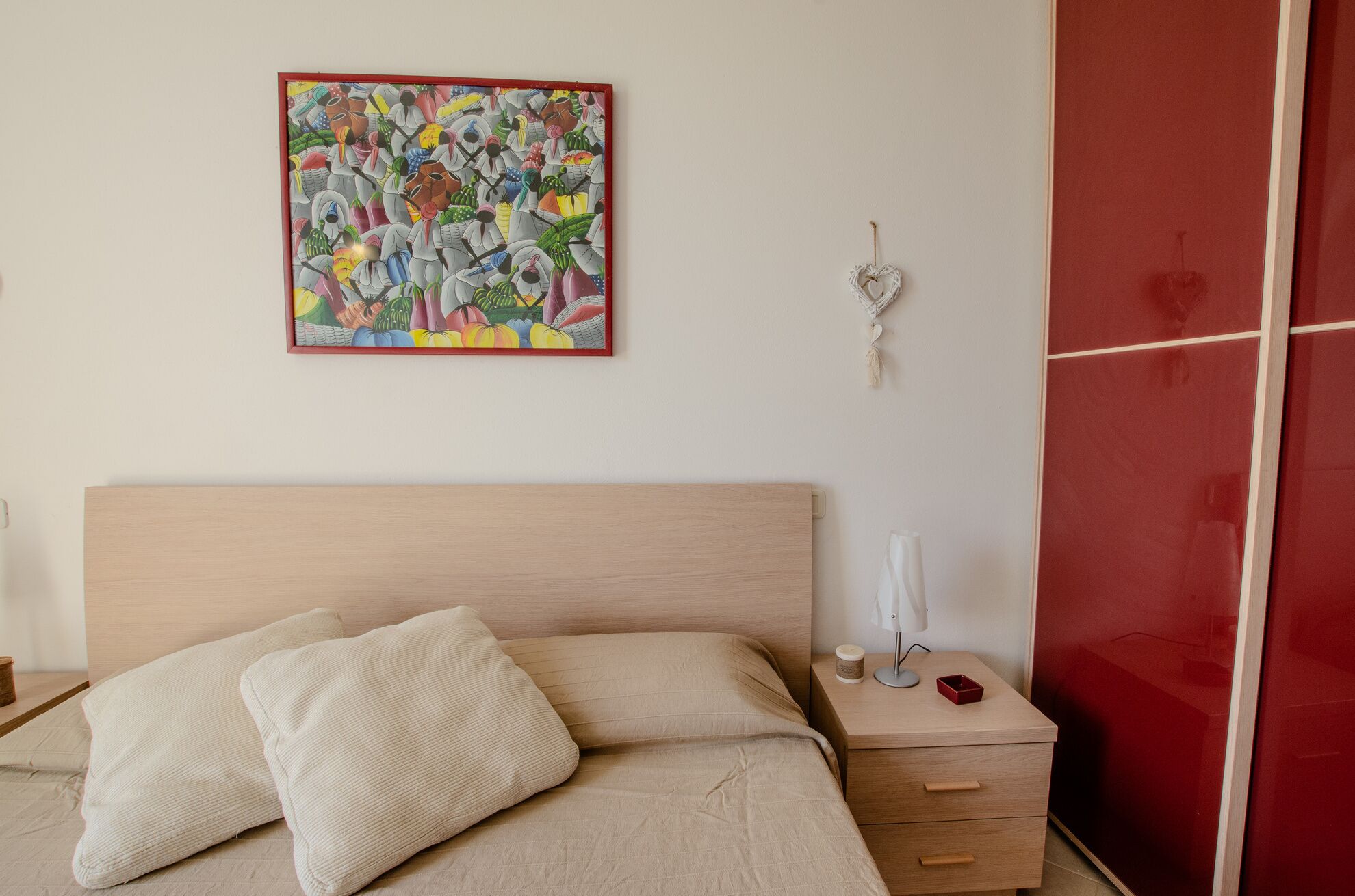 A bedroom with a bed, a nightstand, a red wardrobe, and a colorful abstract painting on the wall.
