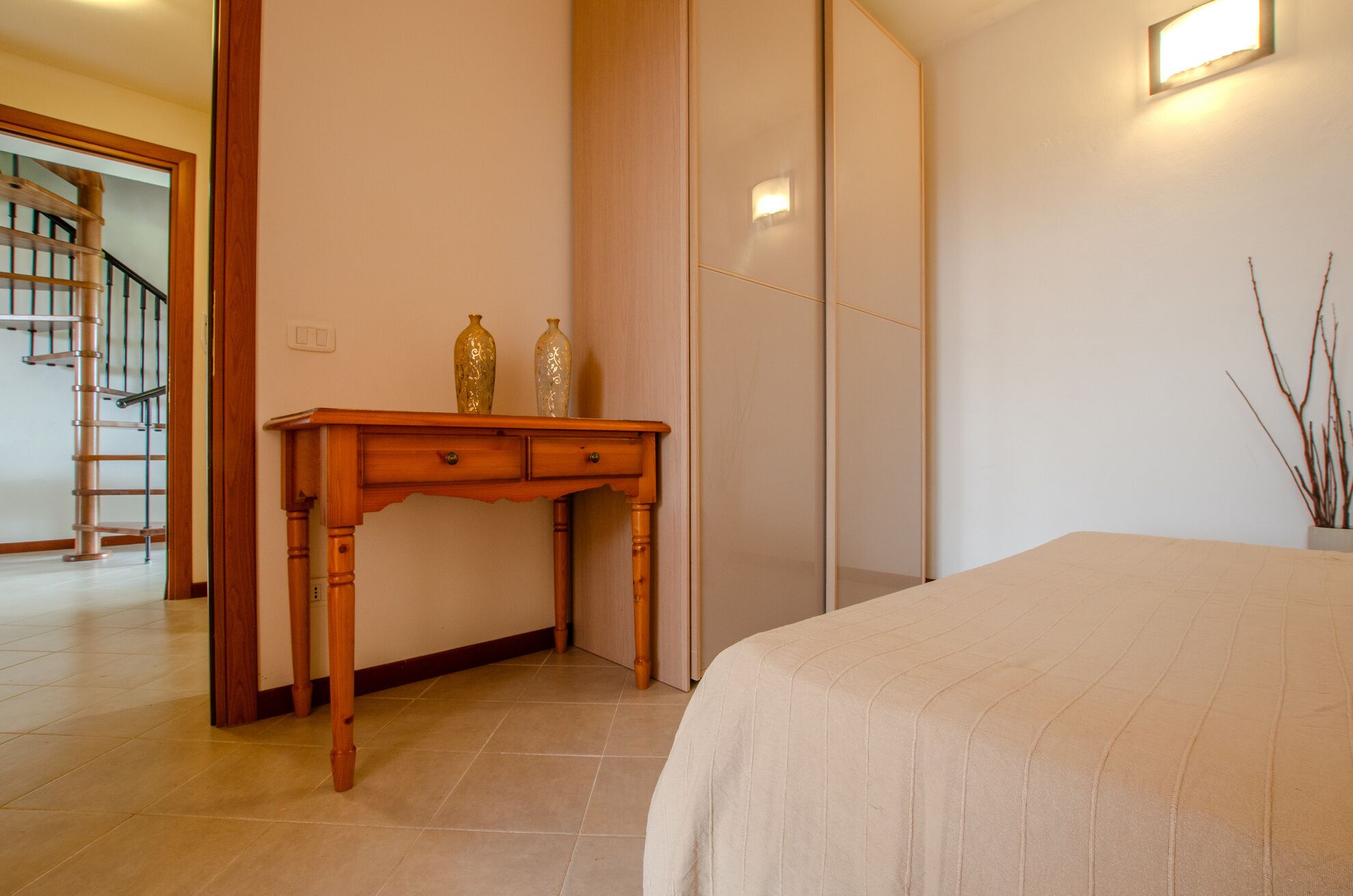 Une chambre à coucher avec un lit, un bureau en bois, deux vases décoratifs, une lampe fixée au mur et un escalier en arrière-plan.