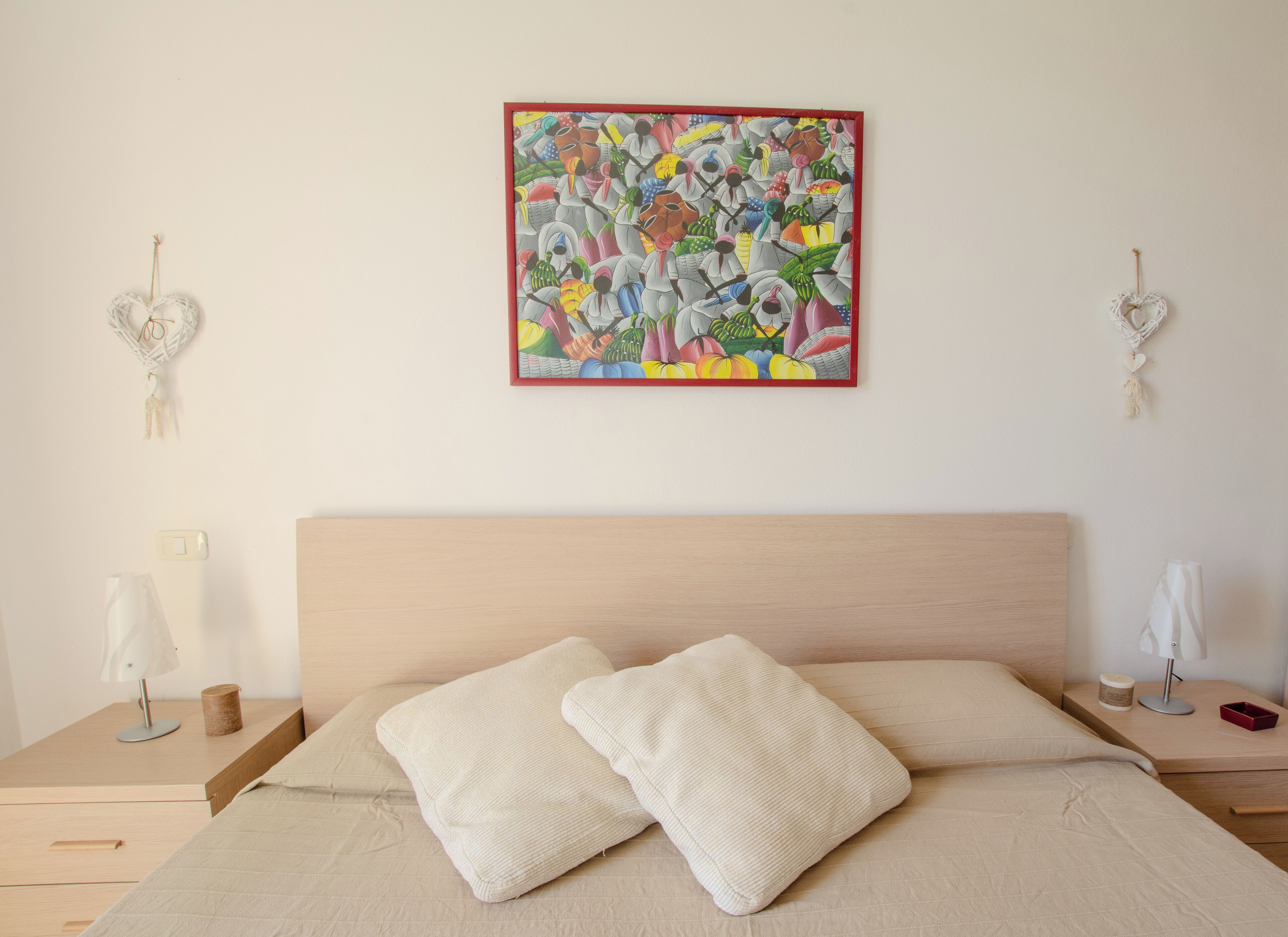 A bedroom with a wooden headboard, a bed with two white pillows, a colorful abstract painting on the wall, and bedside tables with lamps and a small container.