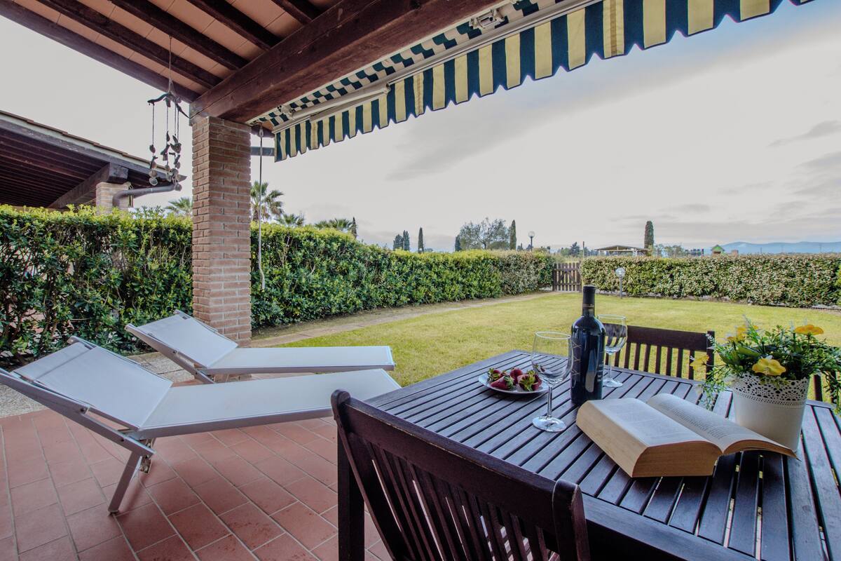 A patio with two lounge chairs, a table with a book, a bottle, and a glass, and a view of a garden and distant landscape.