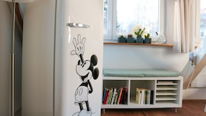 Ein weißer Kühlschrank mit Mickey Mouse Aufkleber, ein Bücherregal mit Büchern, ein Fenster mit Blick auf Bäume und eine Lampe.