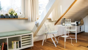 Ein gemütliches Dachzimmer mit schrägen Wänden, einem kleinen Tisch mit Stühlen, einem Bücherregal und einem Fenster mit Pflanzen.