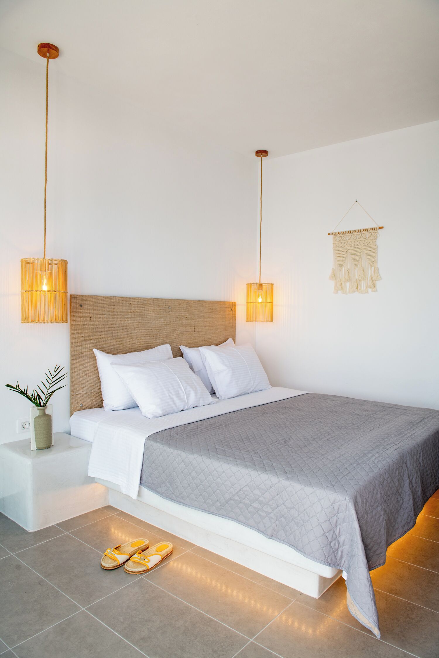 Un dormitorio moderno con una cama grande, lámparas de noche, una planta pequeña y un par de pantuflas en el suelo.