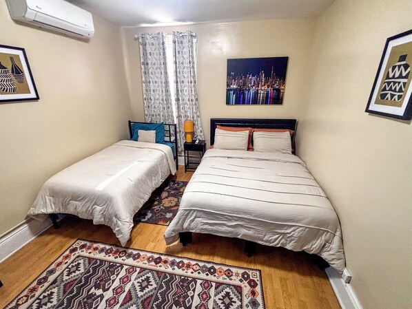 Un dormitorio con dos camas, un tapete estampado, una mesita de noche con una lámpara y un cuadro de un paisaje urbano en la pared.