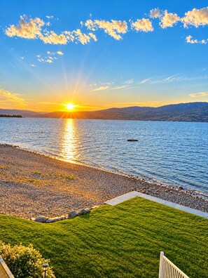 Un coucher de soleil sur un lac paisible, avec une zone herbeuse et un sentier menant à l’eau.