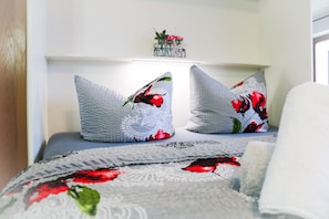 A bed with a floral-patterned blanket, two pillows with a similar design, and a small plant on a shelf above the bed.