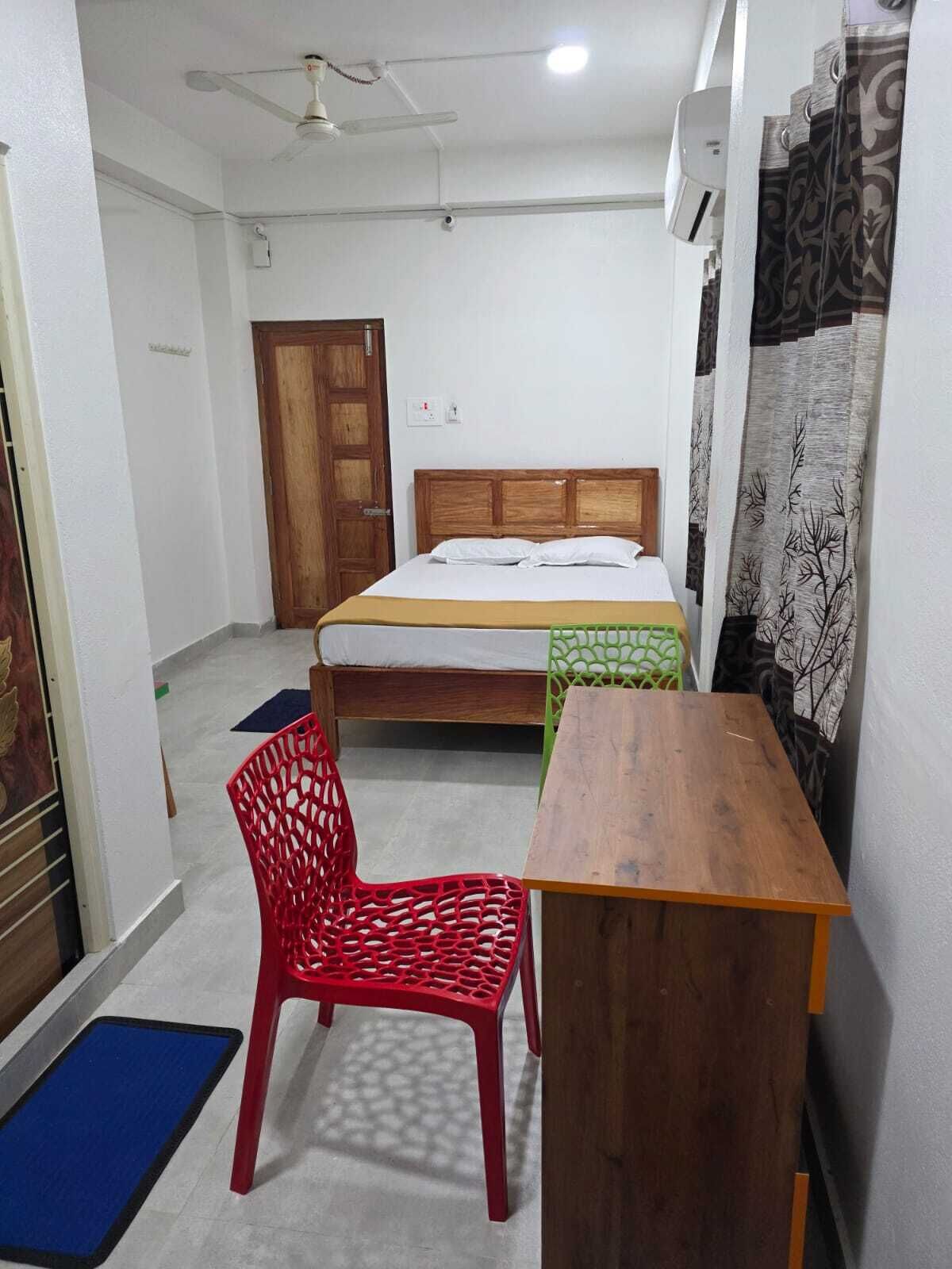 A small, clean room with a single bed, a wooden desk, a red chair, and a ceiling fan.