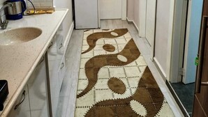 Un pasillo estrecho con una zona de cocina a un lado, que incluye un lavamanos, armarios y una cafetera. Una alfombra estampada cubre el suelo, y se ven dos puertas.